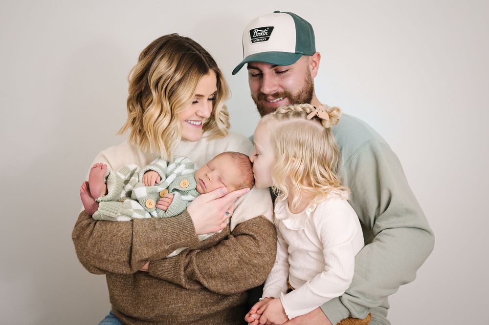 A tender family moment with parents holding their newborn and toddler against a light background.