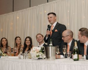 Speaker stands at head table during wedding reception with wedding party seated beside white curtain backdrop.