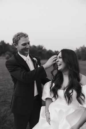 Black and white outdoor wedding photo showing playful moment between a bride and groom in a field.