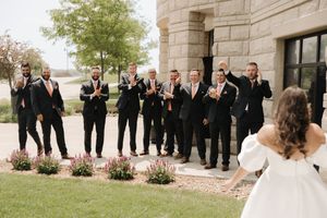 A line of groomsmen in black suits react joyfully outside a stone building during a wedding ceremony.