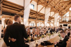 Wedding guests stand and applaud in a rustic reception venue with exposed wooden beams and decorated dining tables.