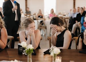 Guests react emotionally during an indoor reception as they sit at tables decorated with candles and white floral centerpieces.