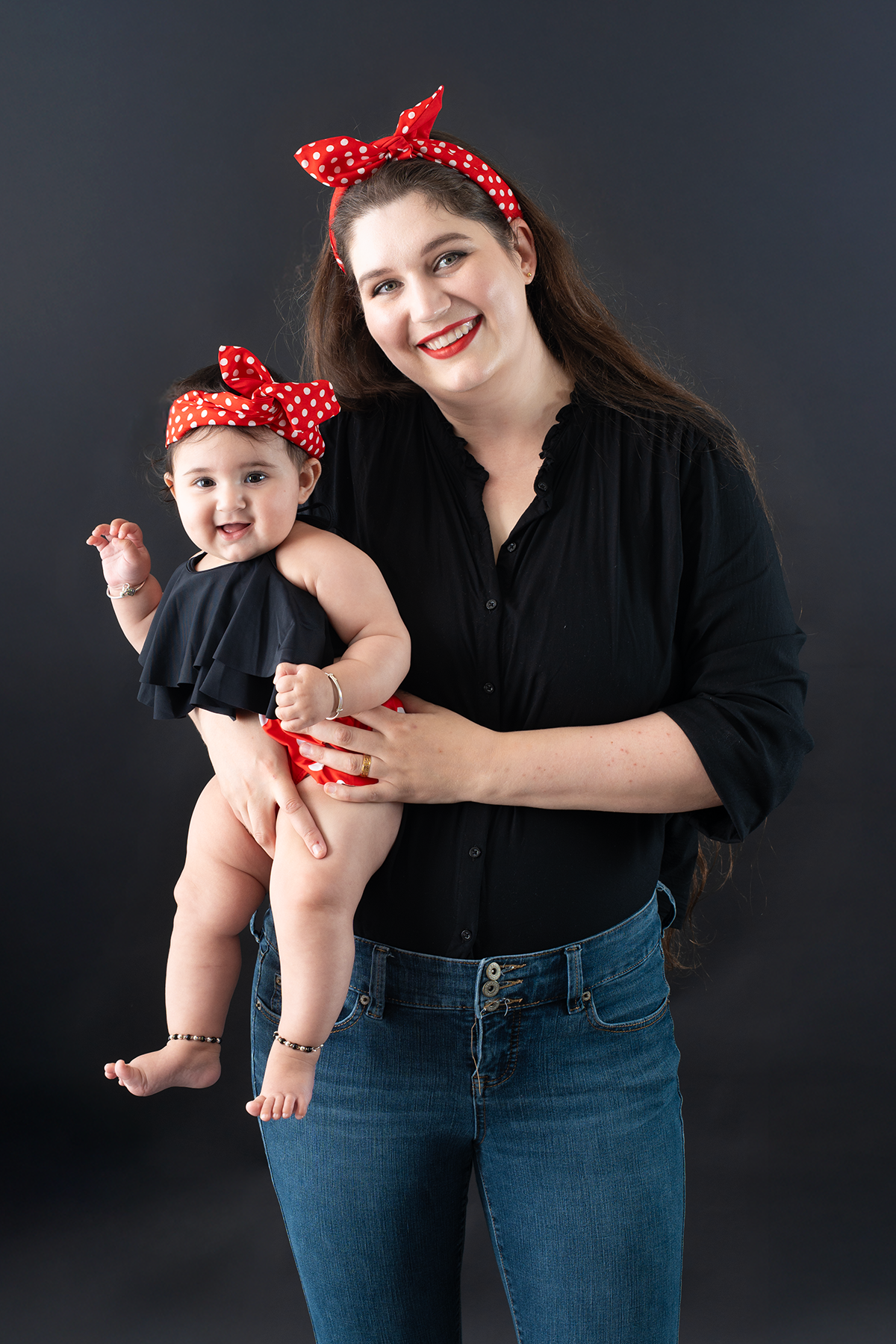 A smiling mother in black shirt and jeans holds her baby girl, both wearing matching red polka dot headbands.