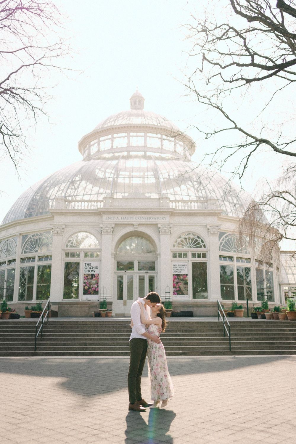 Couple sharing a romantic kiss in front of a grand glass greenhouse conservatory building.