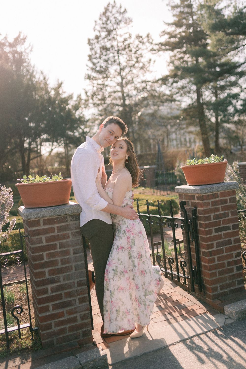 Couple embracing tenderly in golden sunlight beside brick pillars with terracotta planters.