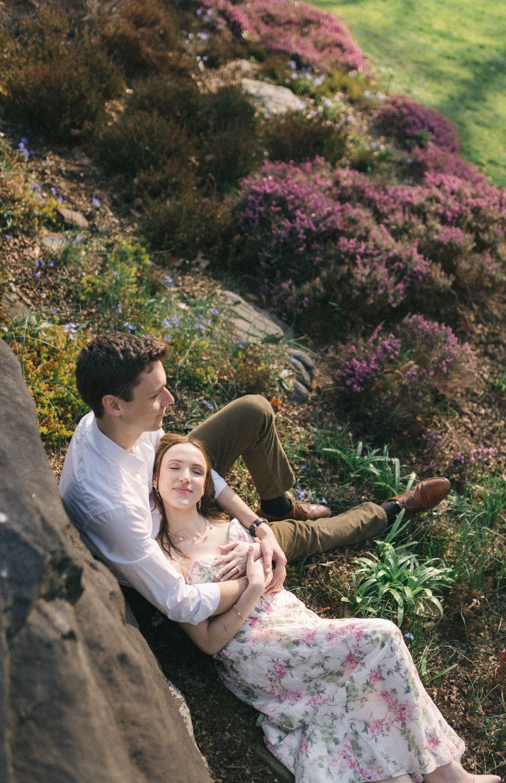 A couple sits together on a rocky hillside surrounded by blooming purple heather, sharing a tender romantic moment outdoors.