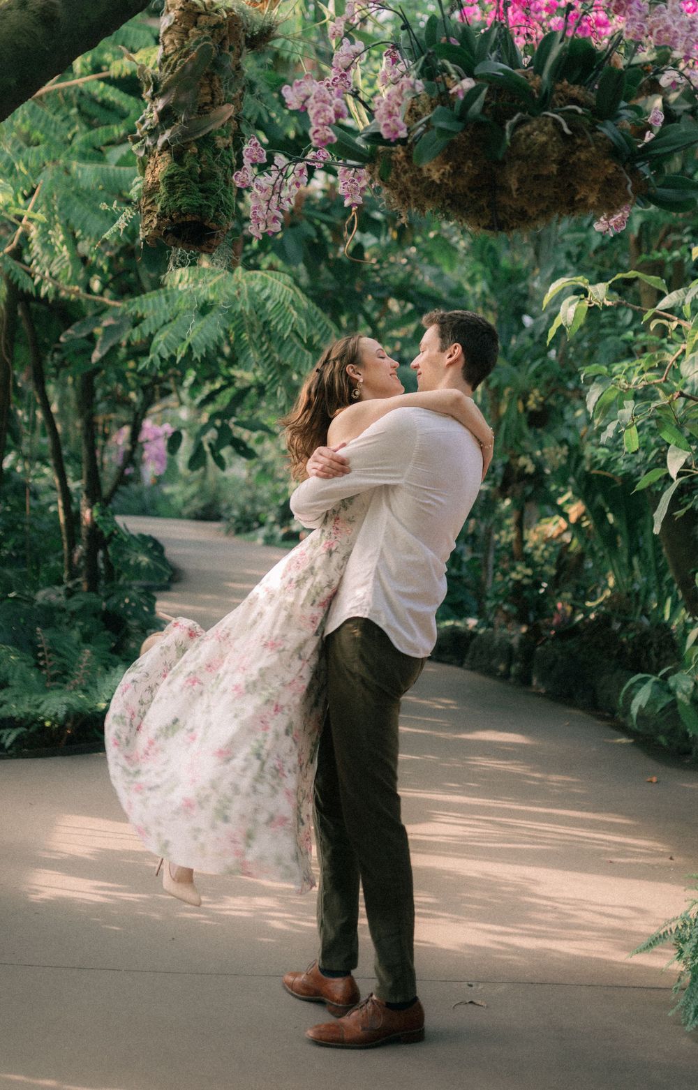 Man lifting woman in floral dress amid lush tropical greenhouse foliage and orchids.