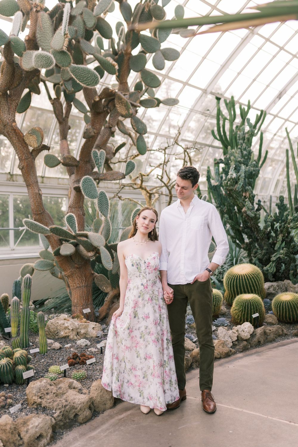 Couple posing in a greenhouse cactus garden, woman in floral dress, man in white shirt and olive pants.