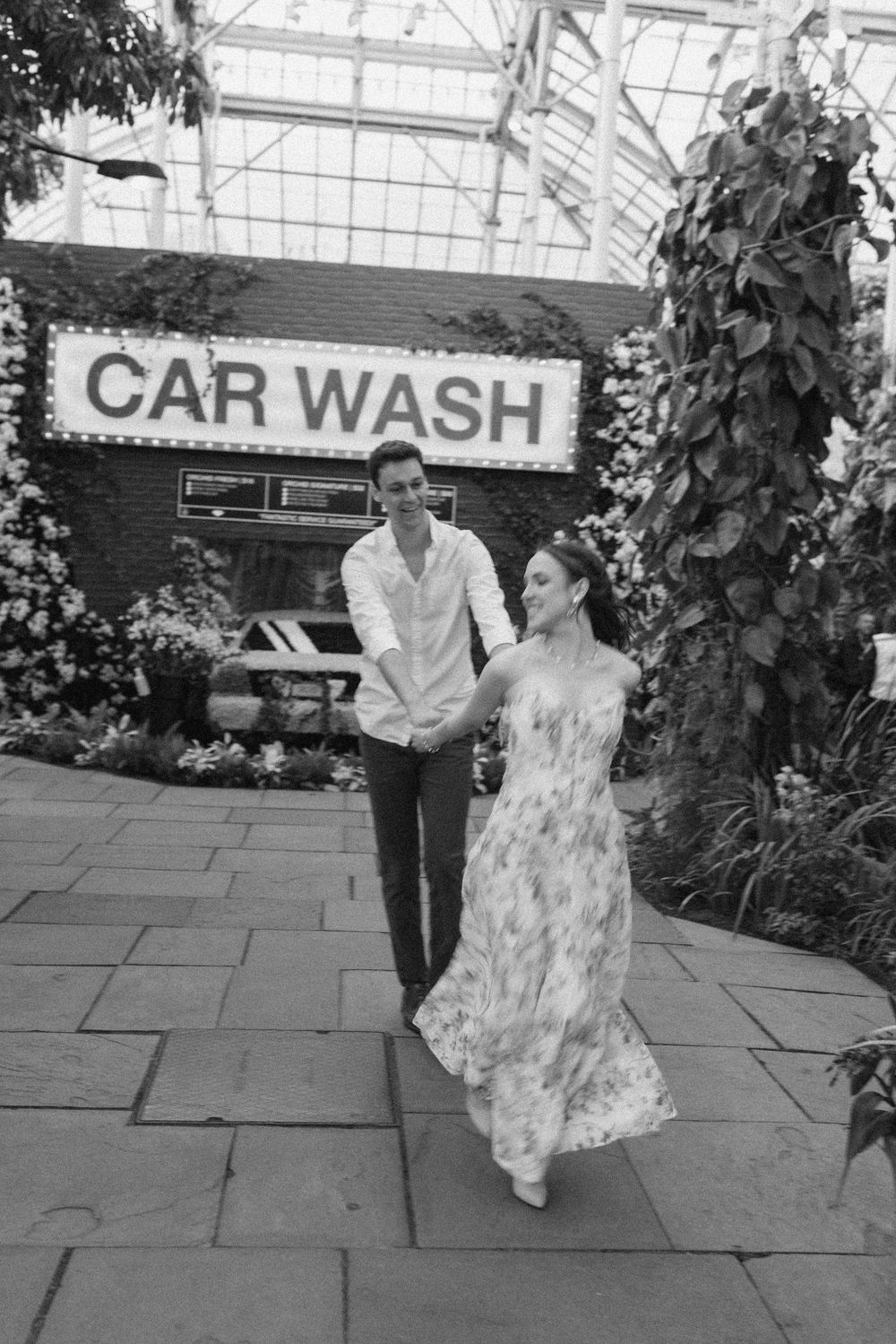 Black and white photo of a couple holding hands and walking past a Car Wash sign surrounded by lush greenery.