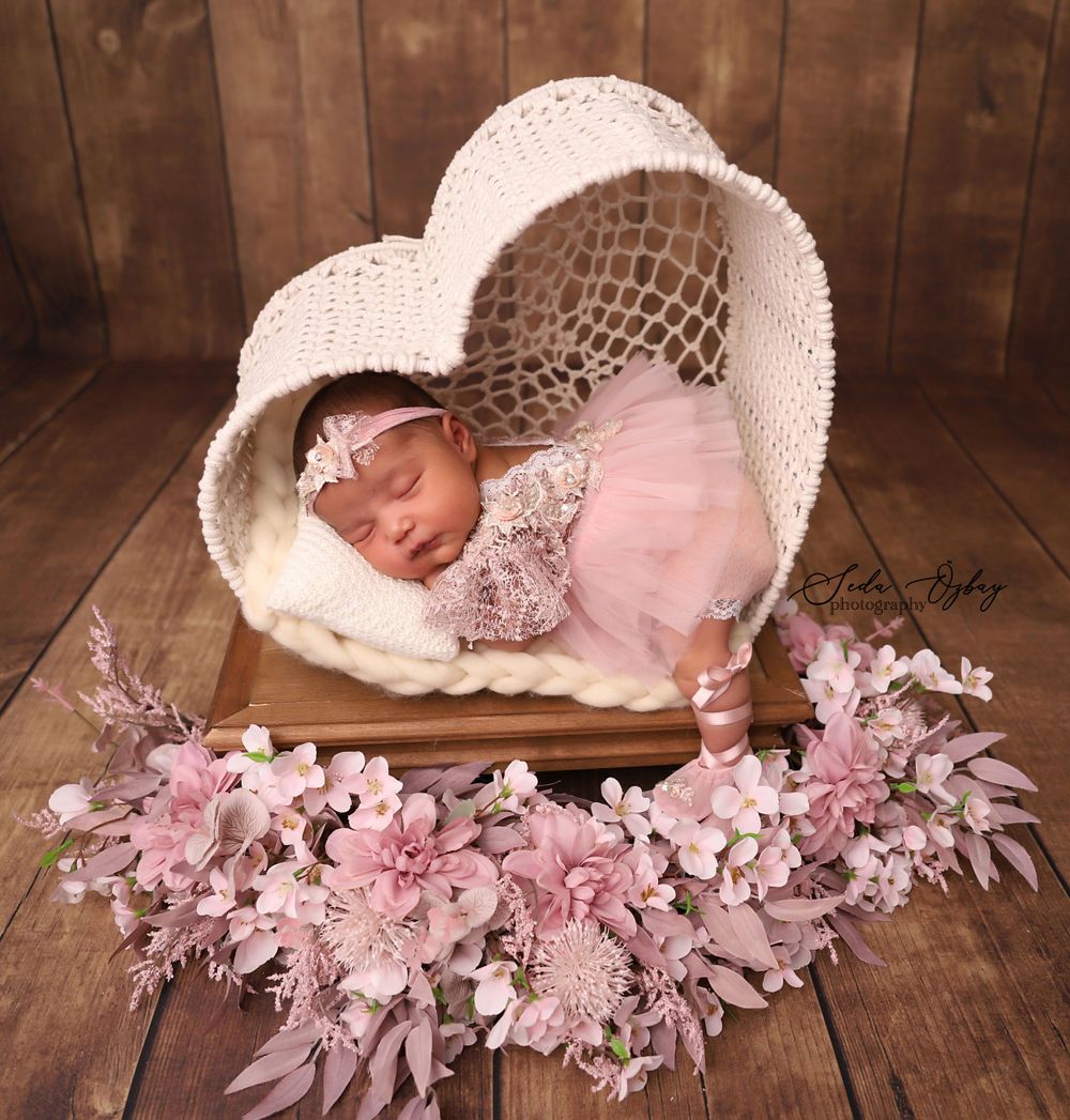 Newborn photography featuring soft pink flowers and white bonnet on rustic wooden backdrop.