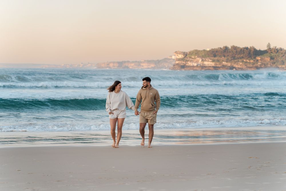 During a Sydney Photoshoot a couple walks hand in hand on a Bondi beach at sunset with turquoise waves and coastal cliffs in the background.