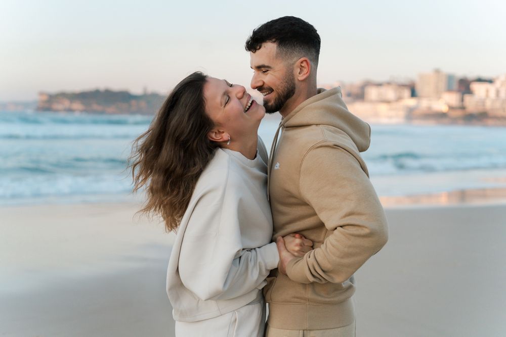 A romantic couple laughing at Bondi beach at sunrise with waves and coastal buildings in the background during a Bondi Beach Photoshoot