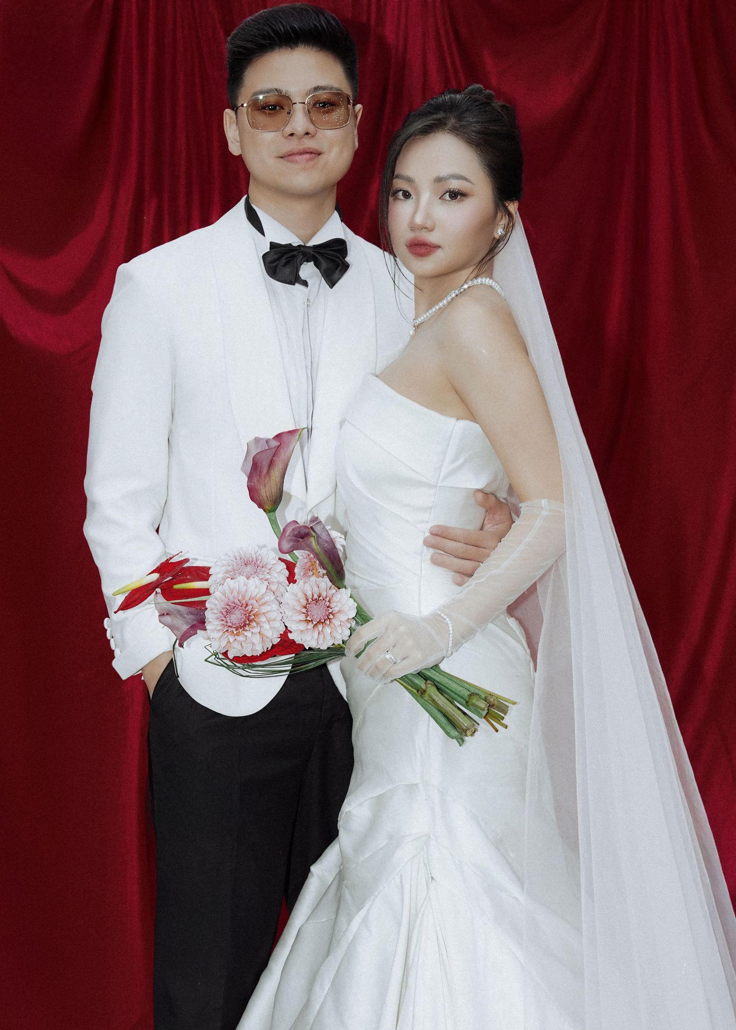 Elegant couple poses for wedding portrait, groom in white tuxedo holding florals, bride in fitted mermaid gown with veil.