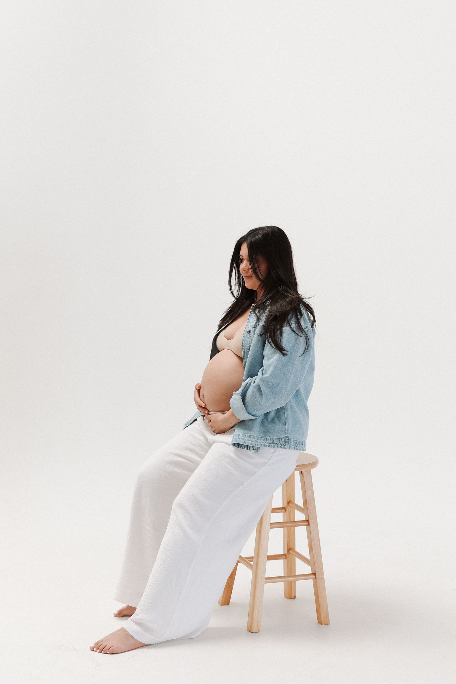Solo Maternity Pictures wearing blue denim jacket and white pants sits on wooden stool against white backdrop