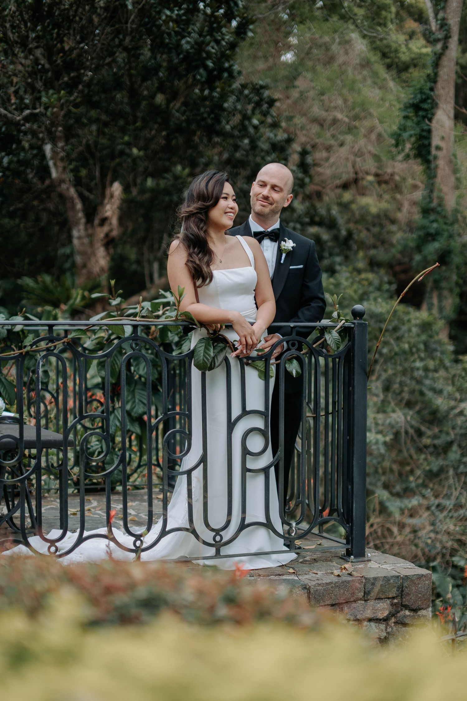 A couple walks together on their wedding day through a garden setting, with the bride in white and groom in black tuxedo.
