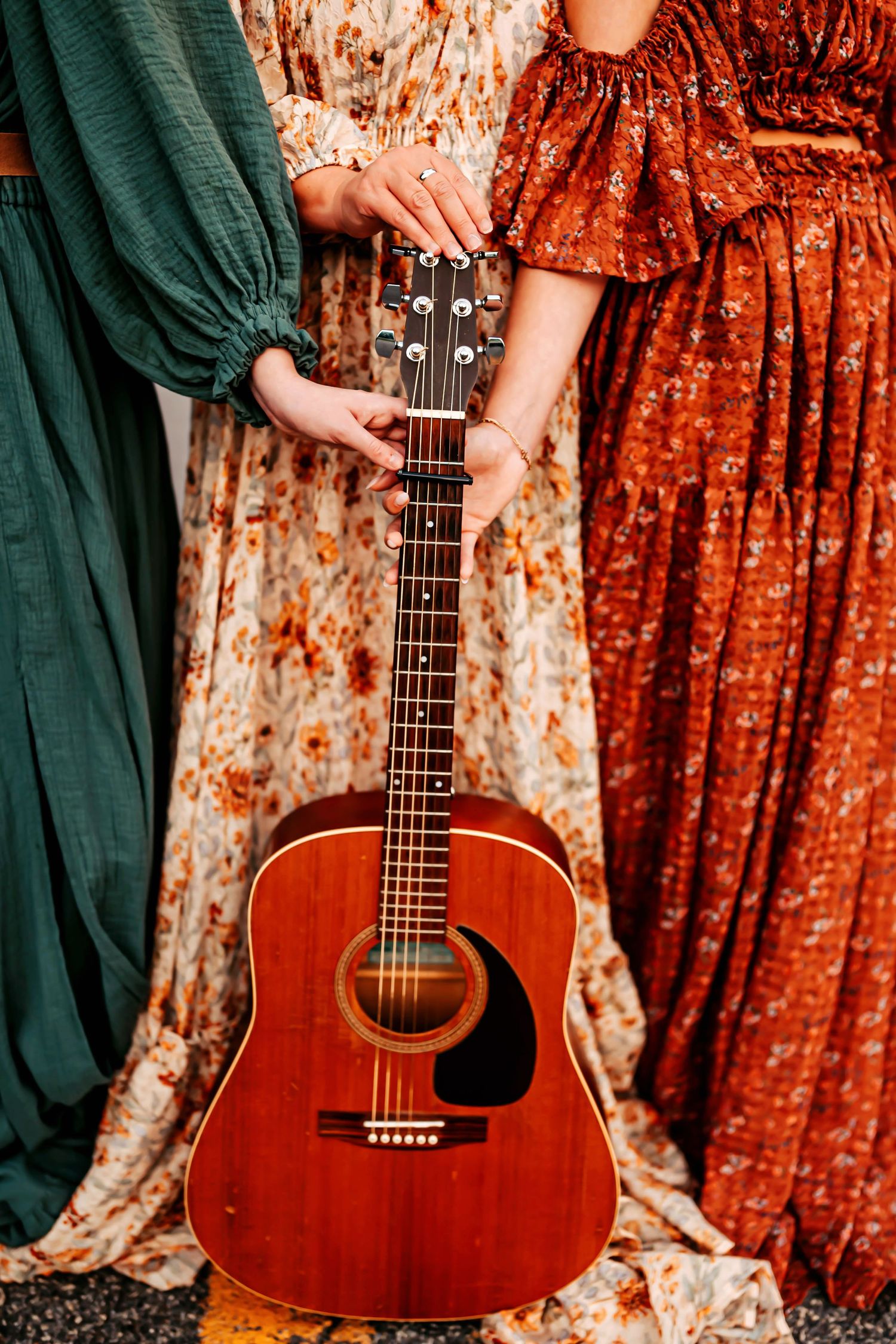 An acoustic guitar stands upright against a backdrop of emerald green and burnt orange vintage dresses.