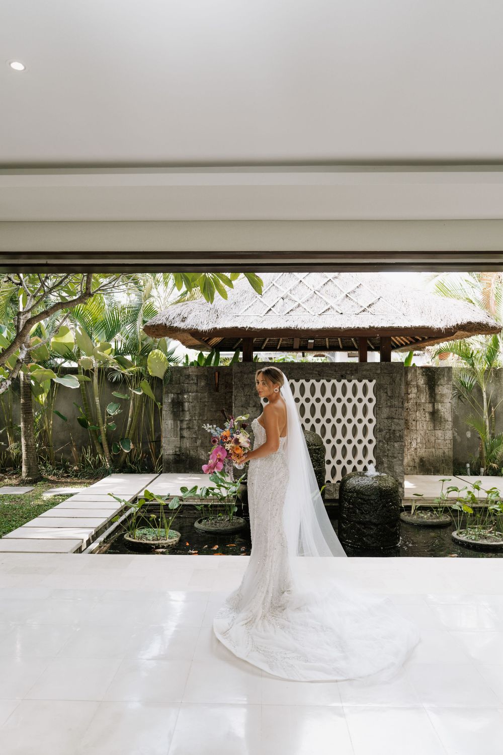 Elegant wedding gown with long flowing train and veil photographed outdoors near a stone wall and tropical greenery.