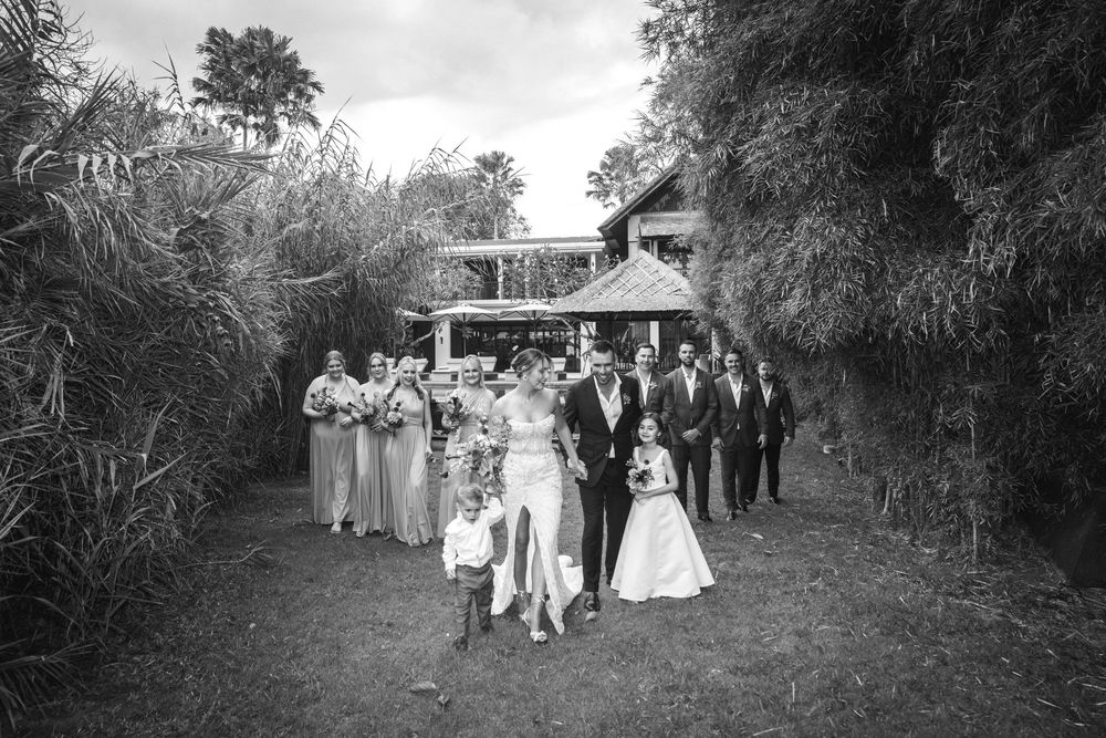 Black and white photograph of wedding party walking down an outdoor path lined with trees toward a venue.