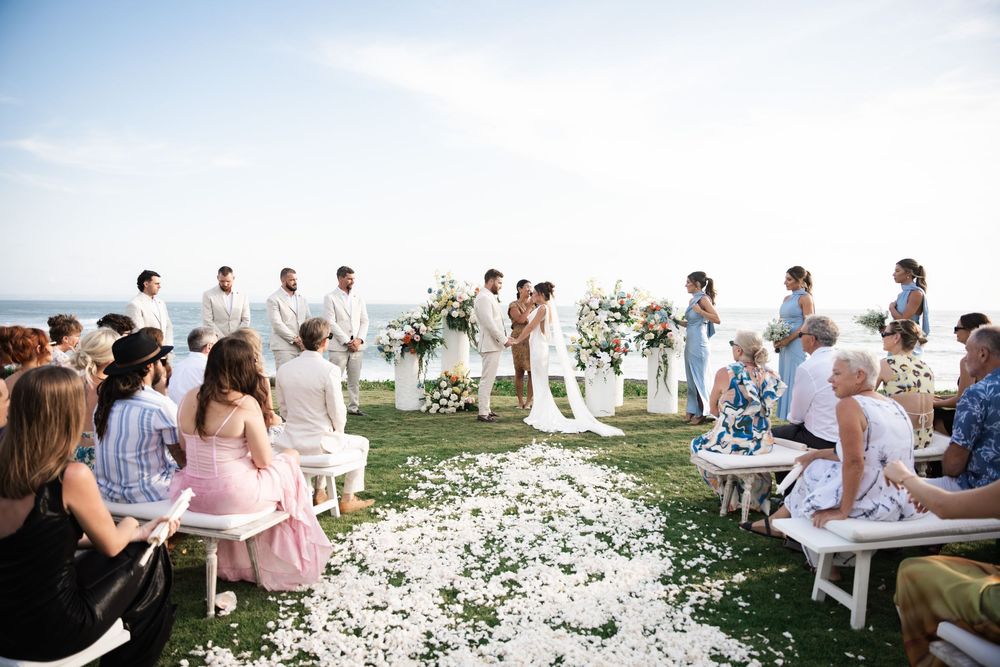 Wedding ceremony guests seated on white chairs with petals scattered along the aisle during an outdoor celebration.
