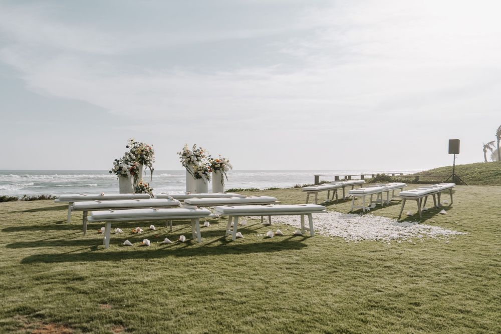 Rustic wooden benches and chairs line up on a grassy beachfront setting for an elegant outdoor wedding ceremony.