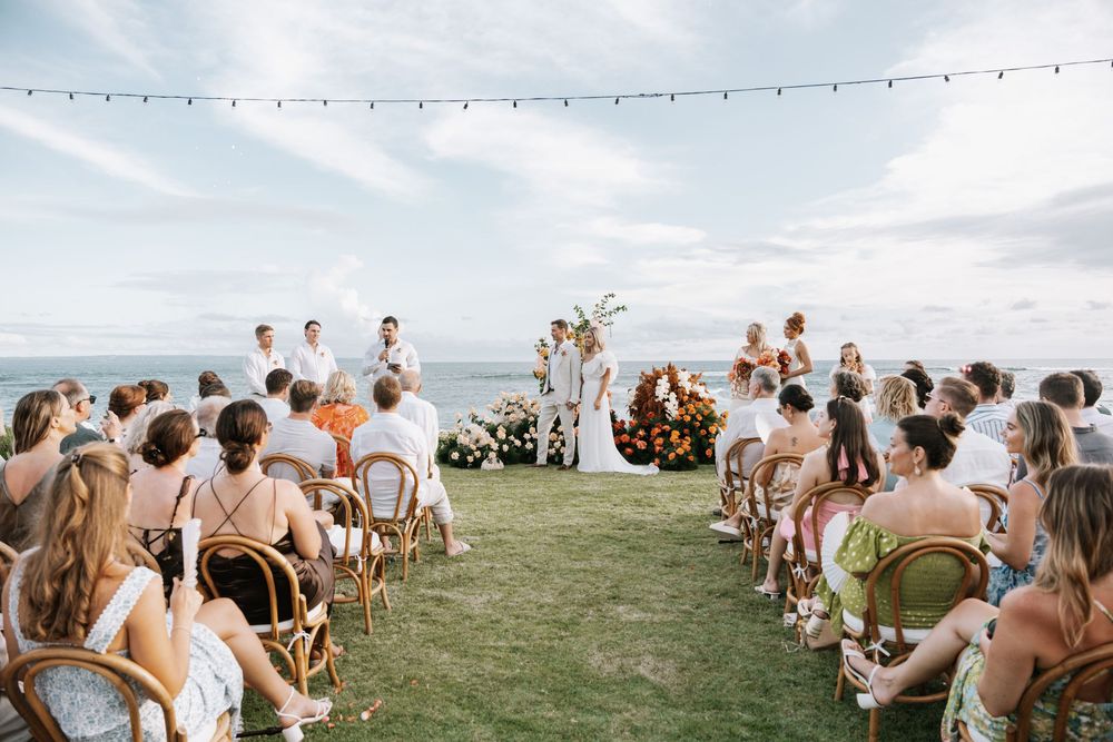 Wedding guests gathered on grass for a scenic outdoor ceremony under string lights during golden hour.