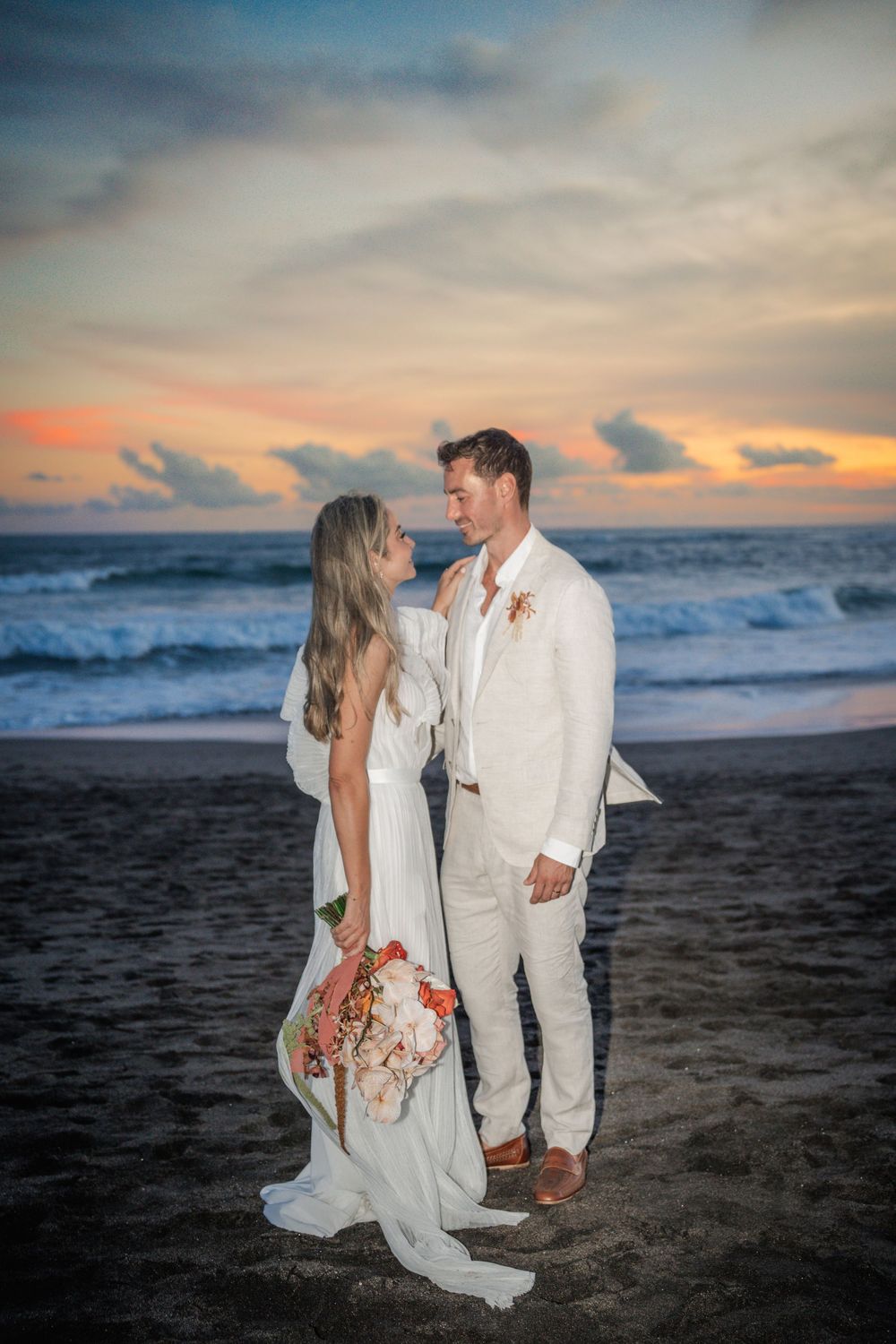 Couple in elegant white wedding attire poses on a sandy beach at sunset with ocean waves in the background.