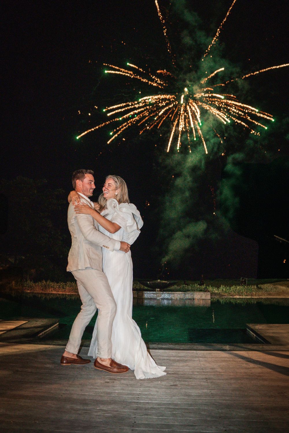 A couple in white attire dances on a wooden deck at night while fireworks burst in the dark sky above them.