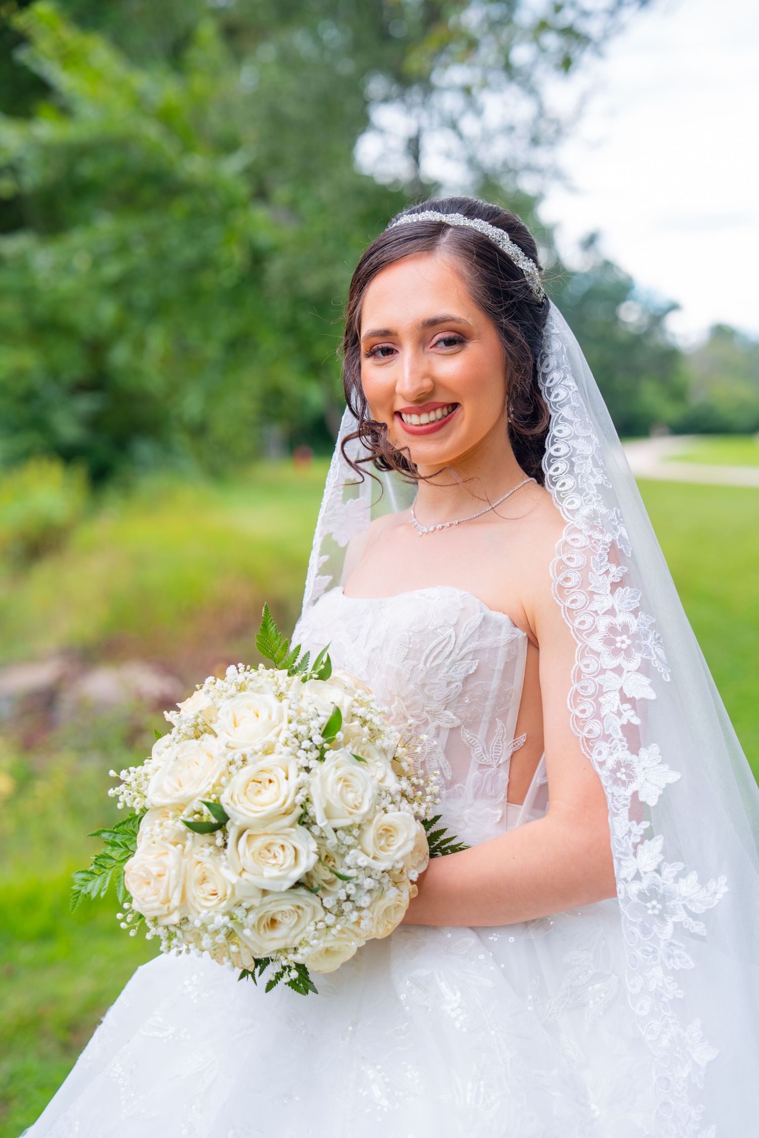 Bride holds elegant white rose bouquet while wearing lace veil outdoors.