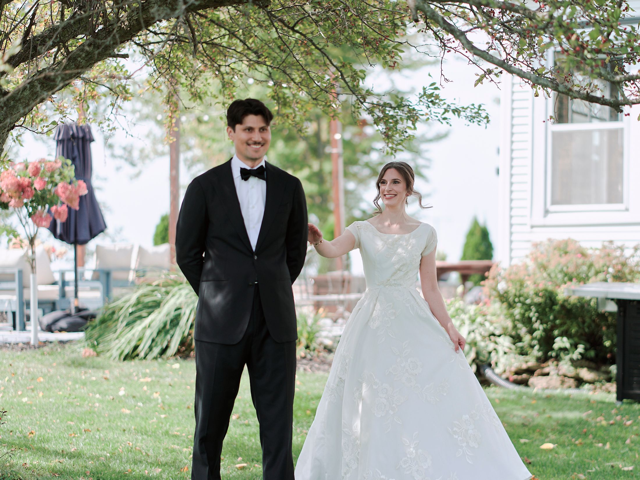 Romantic wedding couple poses together outdoors on a grassy lawn surrounded by trees.