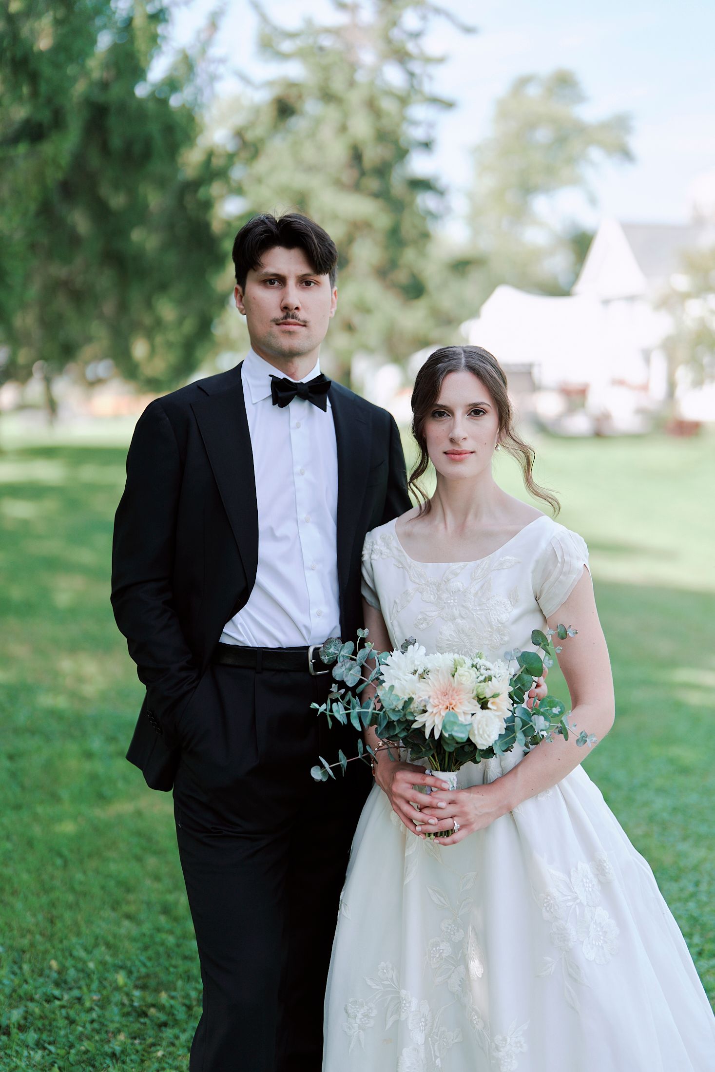 A couple in formal wedding attire poses together on a grassy lawn during a sunny outdoor photo session.