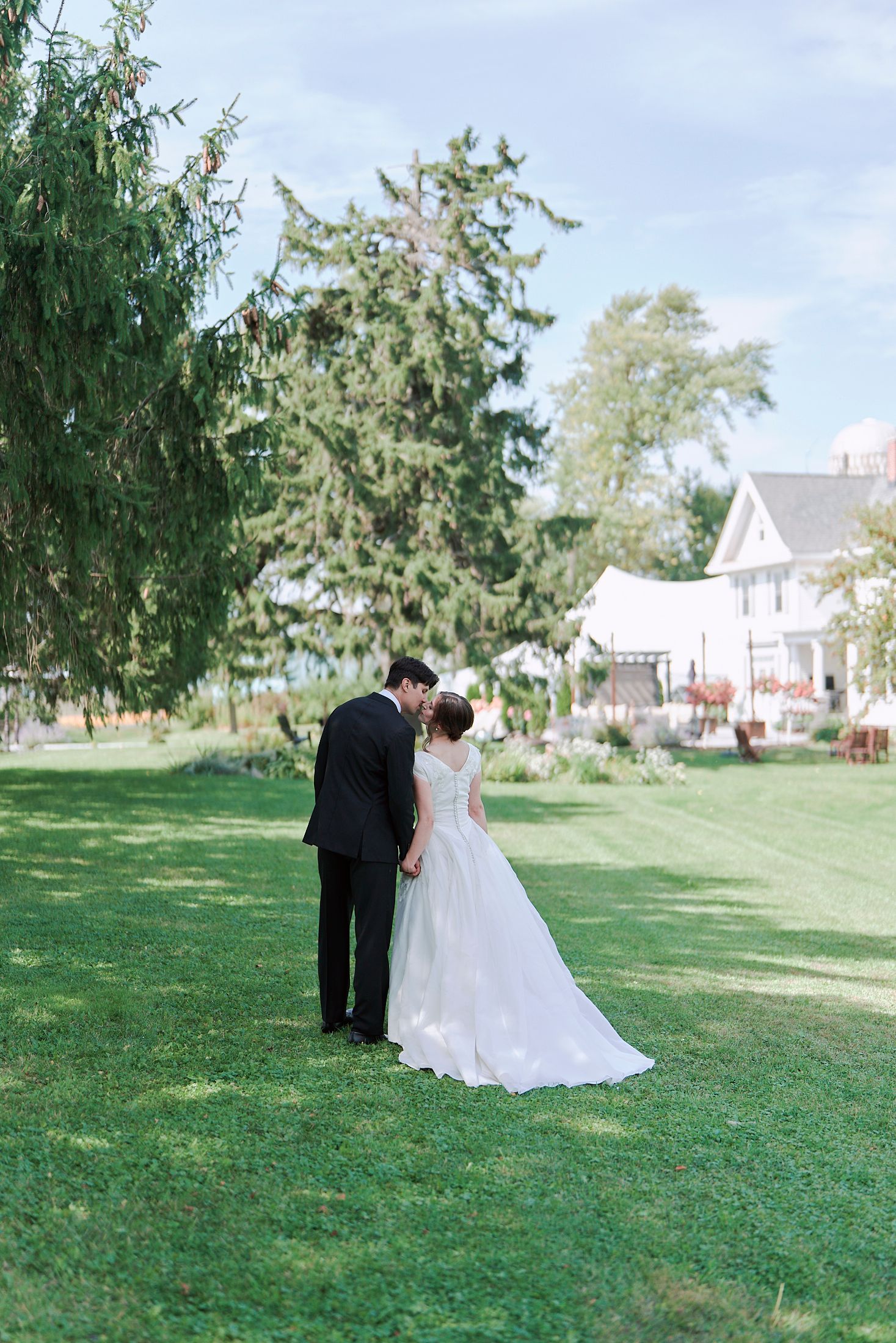 A romantic wedding moment captured on a lush green lawn with tall evergreens and white buildings in the distance.
