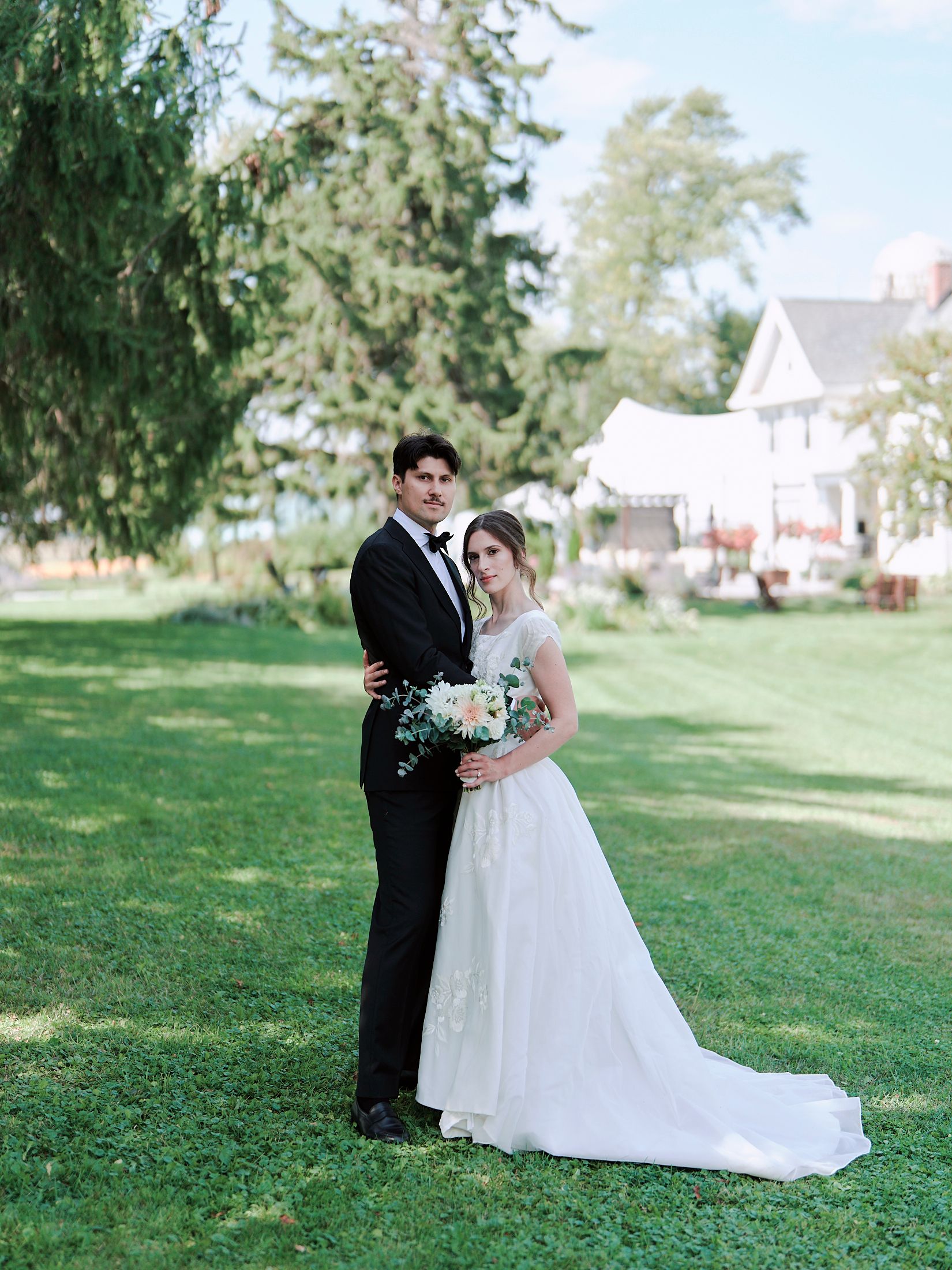 A couple in formal wedding attire embrace on a grassy lawn with trees and houses in the background.
