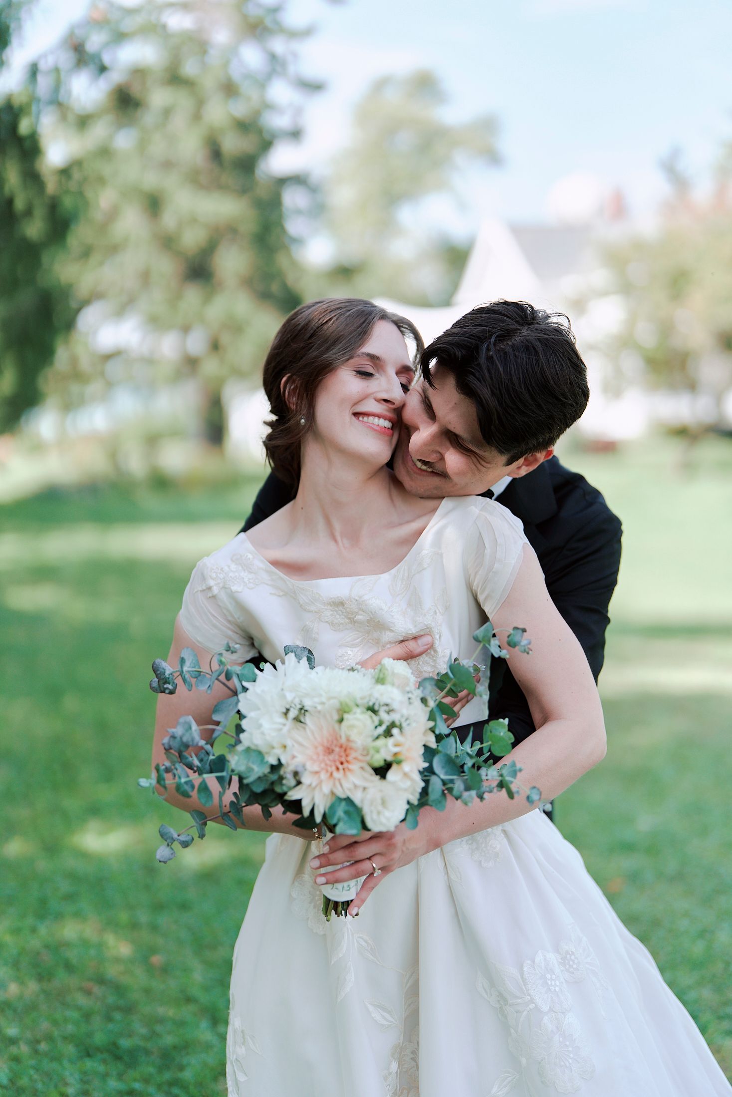 A romantic wedding couple embraces outdoors while holding a delicate bouquet of white flowers and eucalyptus.