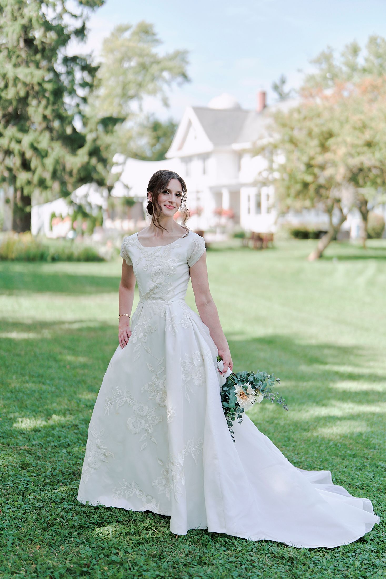 A bride in a classic white lace wedding gown poses gracefully on a sunny lawn with houses in the background.