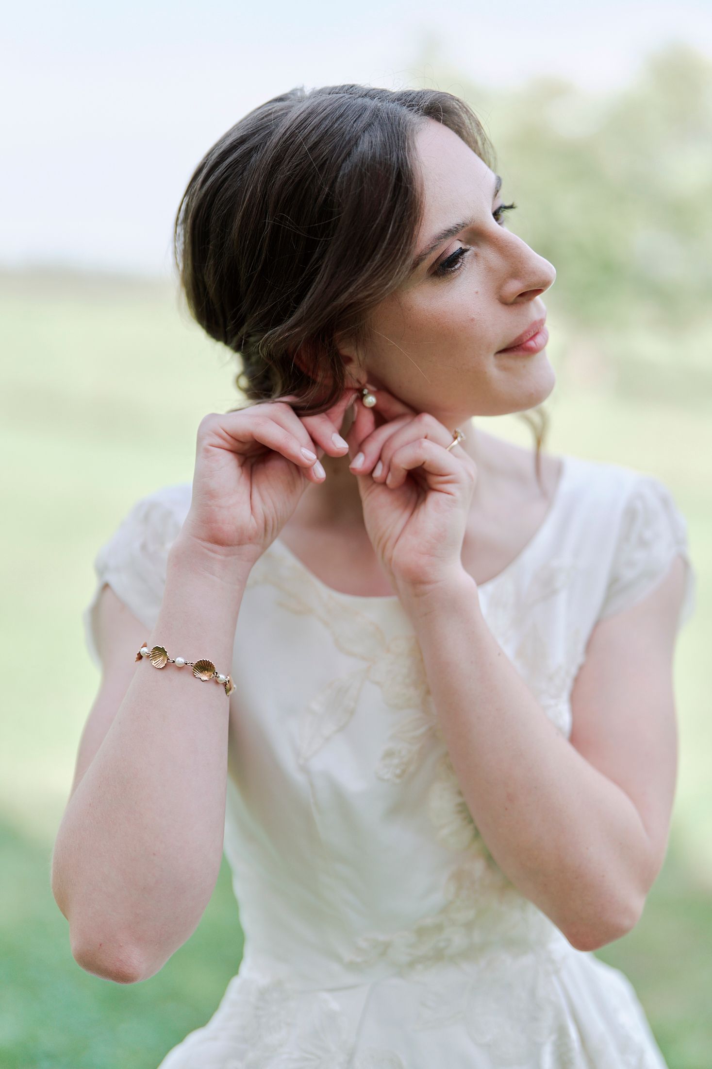 A person in a white dress poses outdoors while adjusting their earrings against a soft blurred natural background.