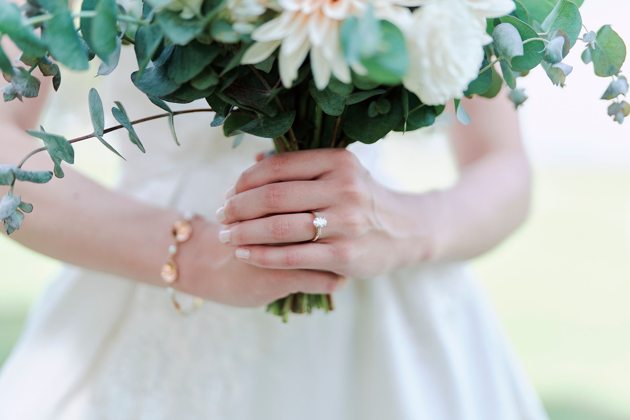 Close-up of hands holding white wedding bouquet with eucalyptus, showing delicate bracelet and engagement ring.