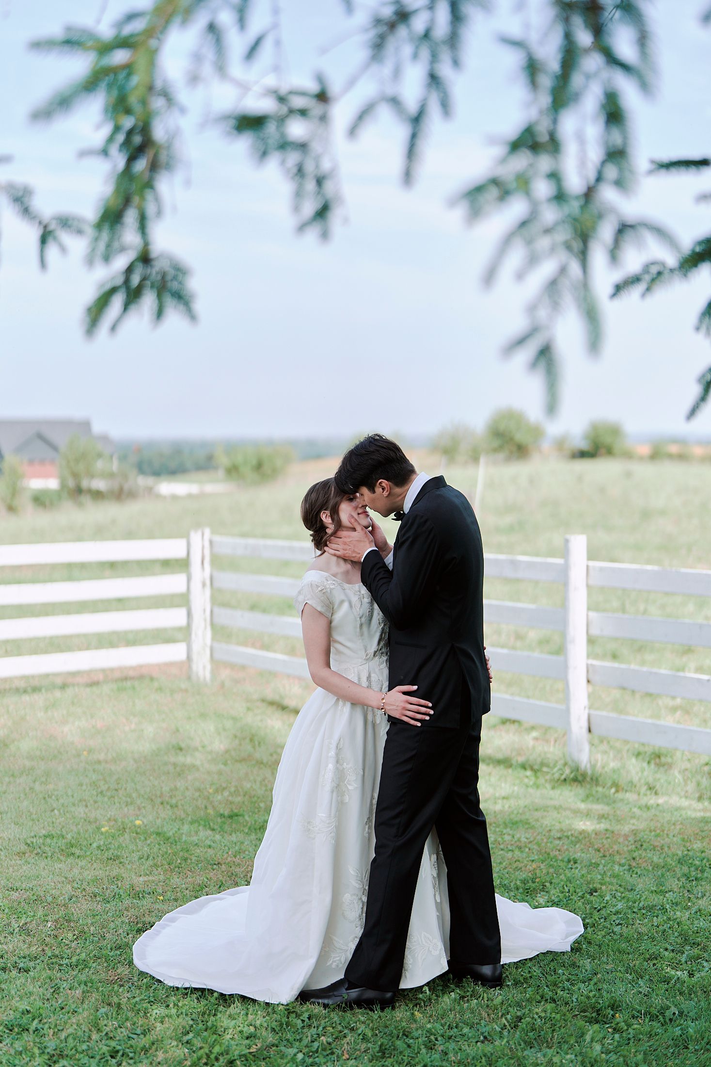 A romantic wedding couple embraces on a grassy hillside with white fencing in the background.