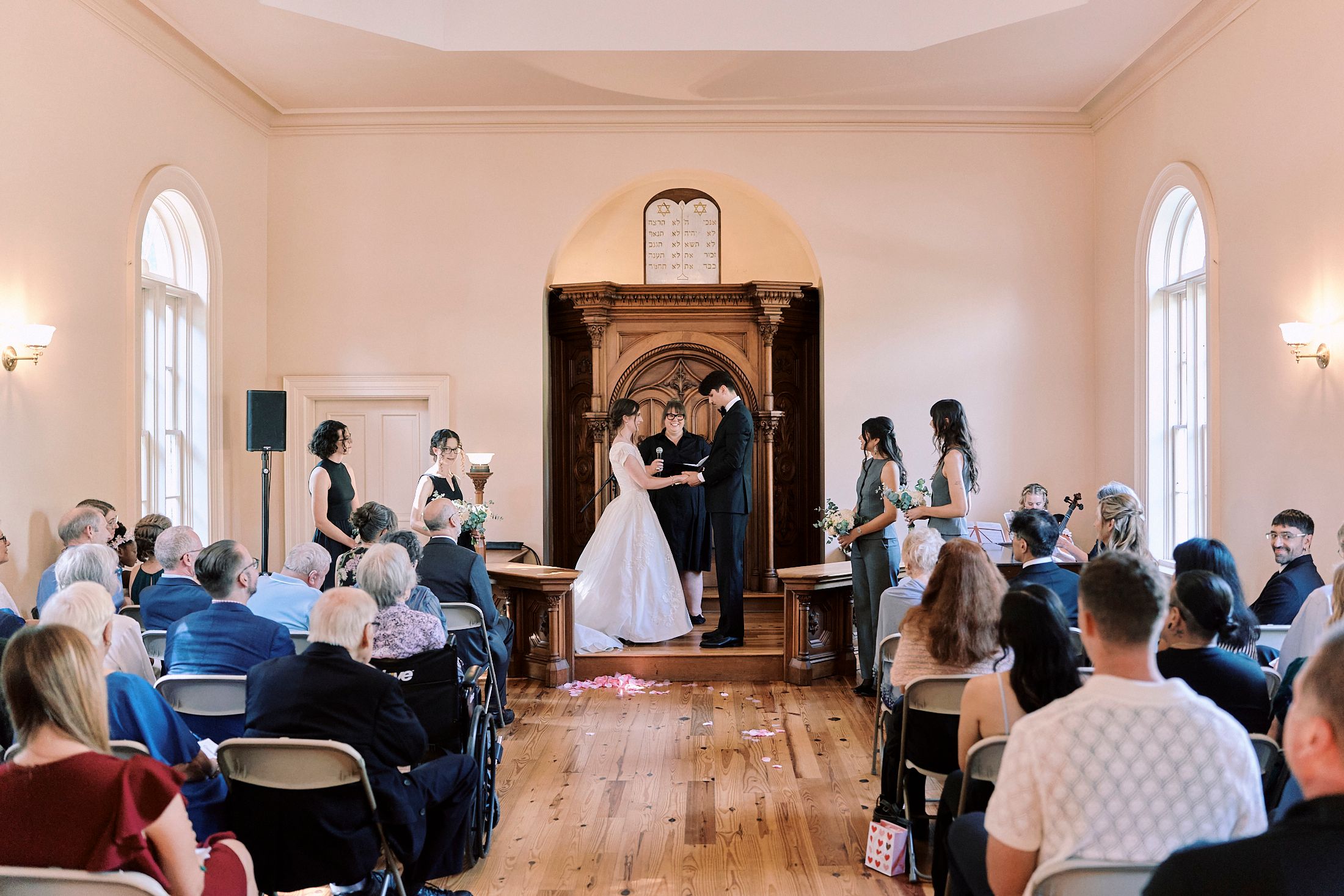 Wedding guests sit in rows of chairs inside a bright, elegant chapel with arched windows during a ceremony.