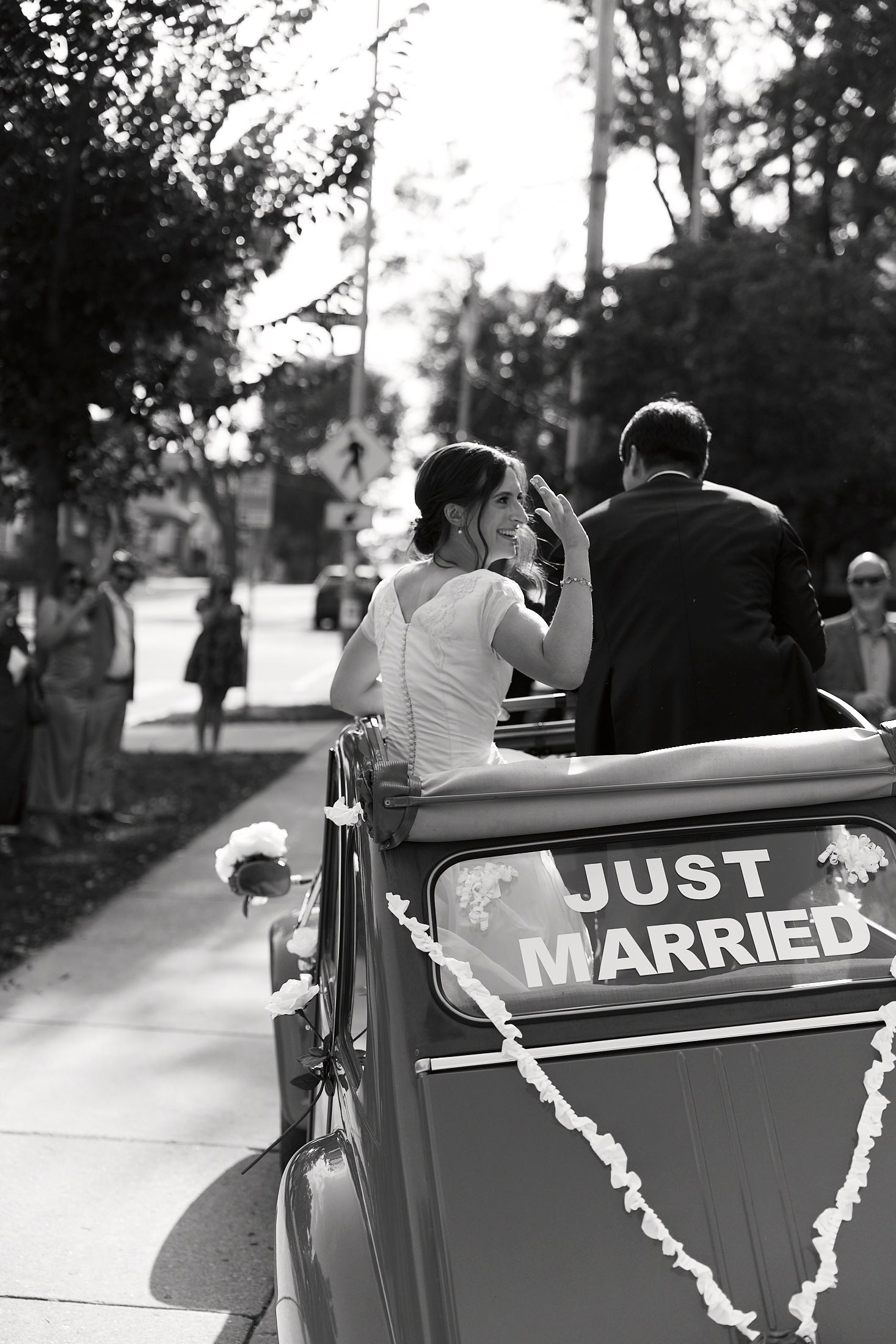 Classic car decorated with 'Just Married' sign and white ribbons parked on a tree-lined road.