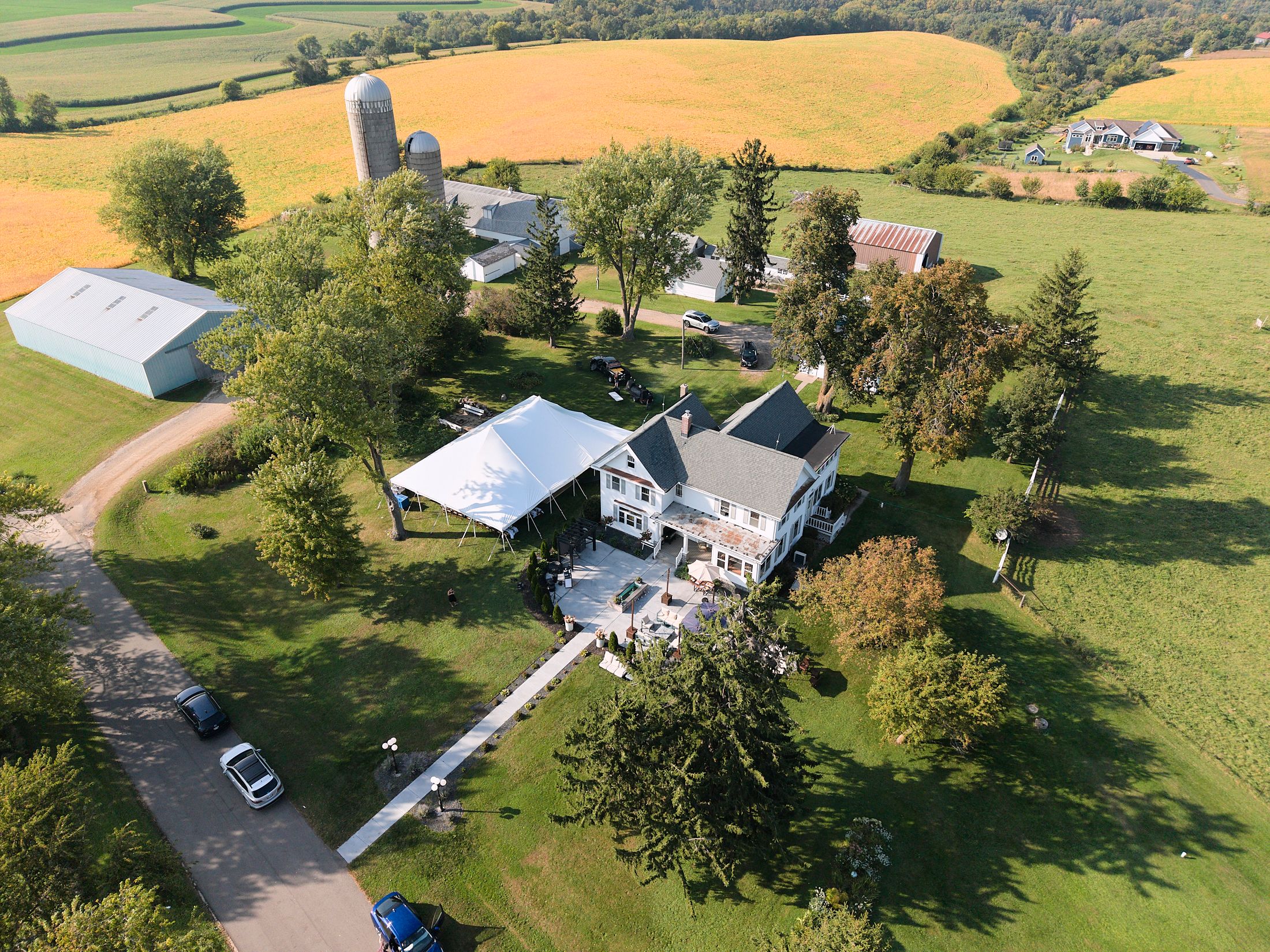 Aerial drone footage shows a white farmhouse and outbuildings nestled among green fields and trees.