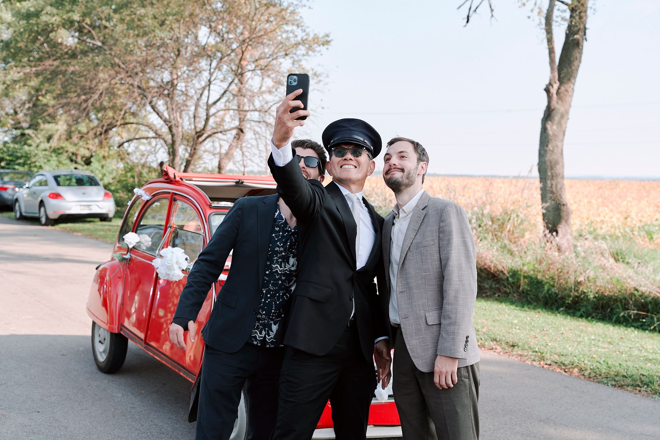 Three people stand together outdoors on a paved path with autumn trees and a red car in the background.