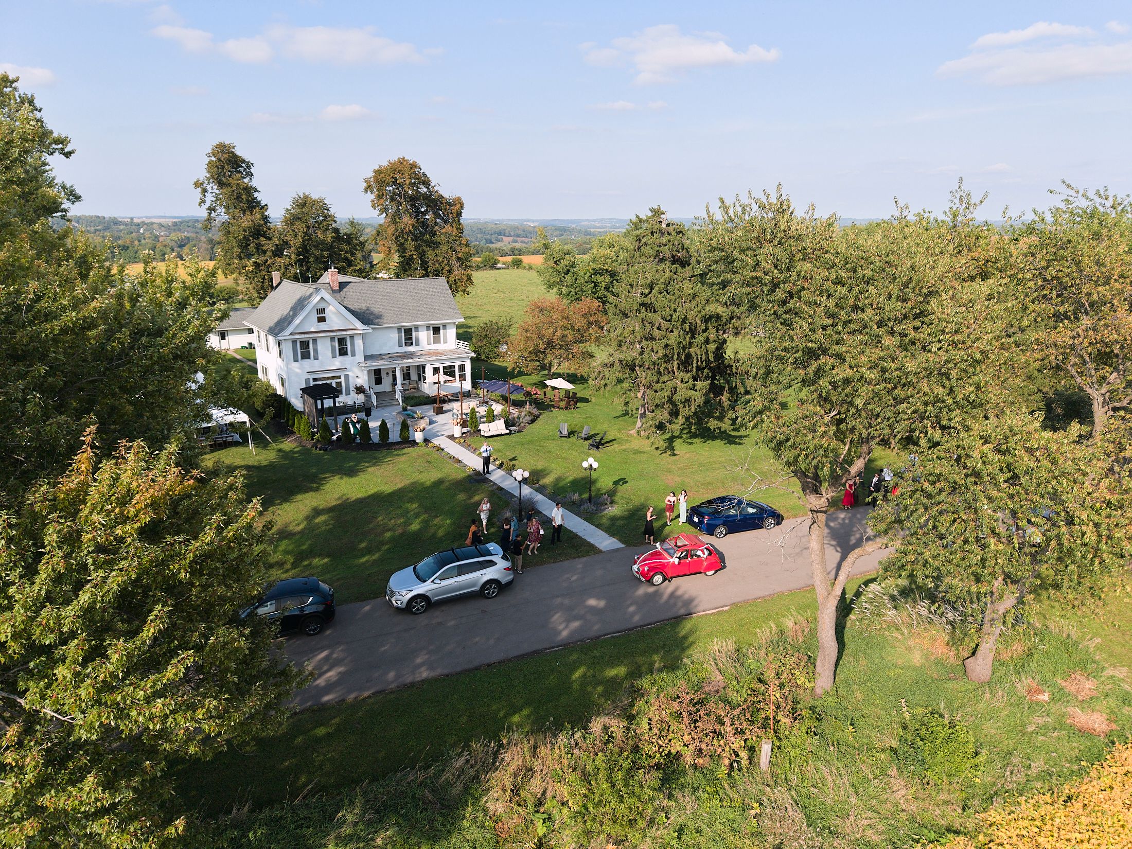 Aerial panoramic view of a rural property with white farmhouse and winding driveway surrounded by green landscape.