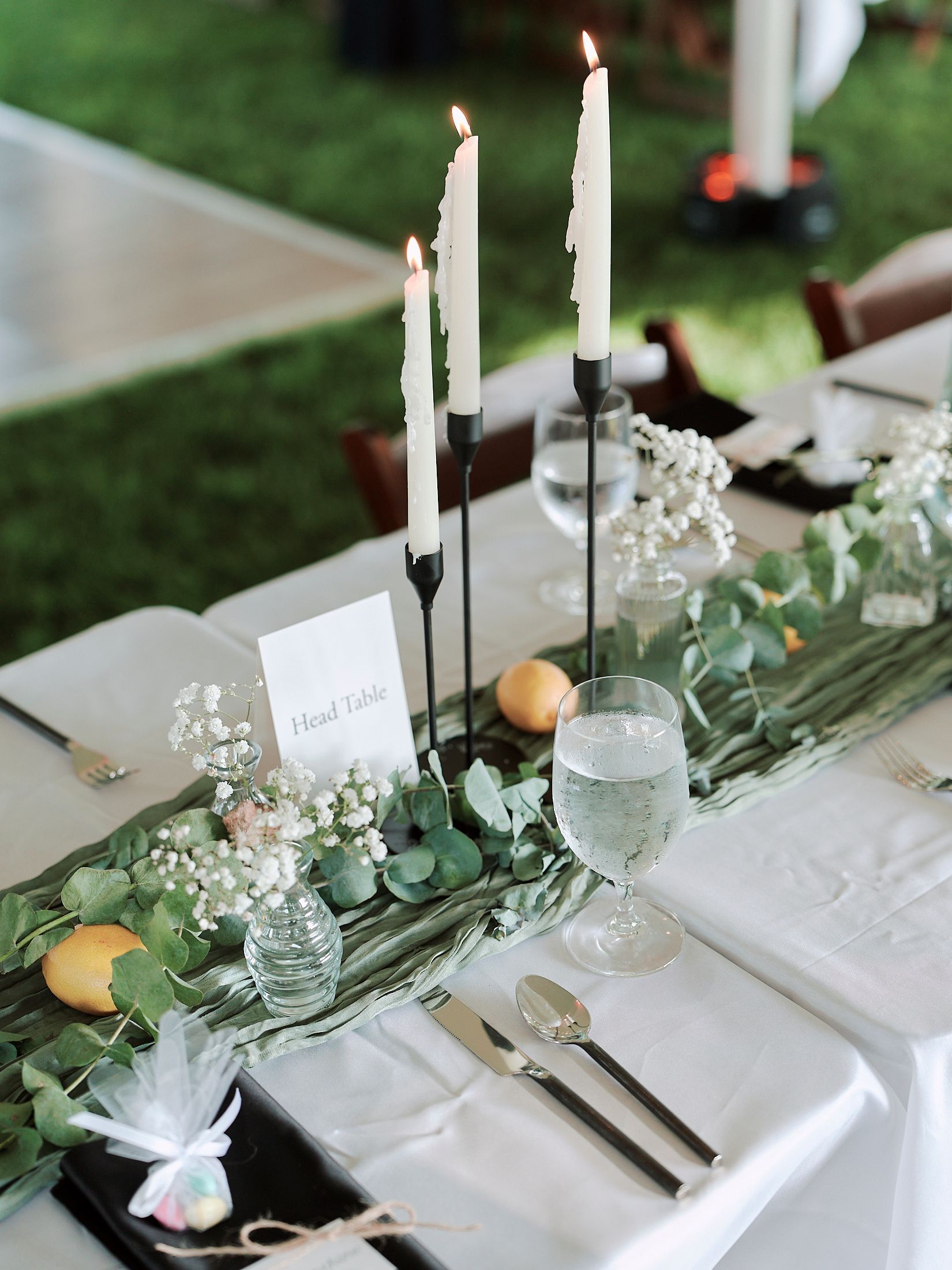 Elegant table setting with white taper candles, citrus fruits, and delicate floral arrangements on a white tablecloth.