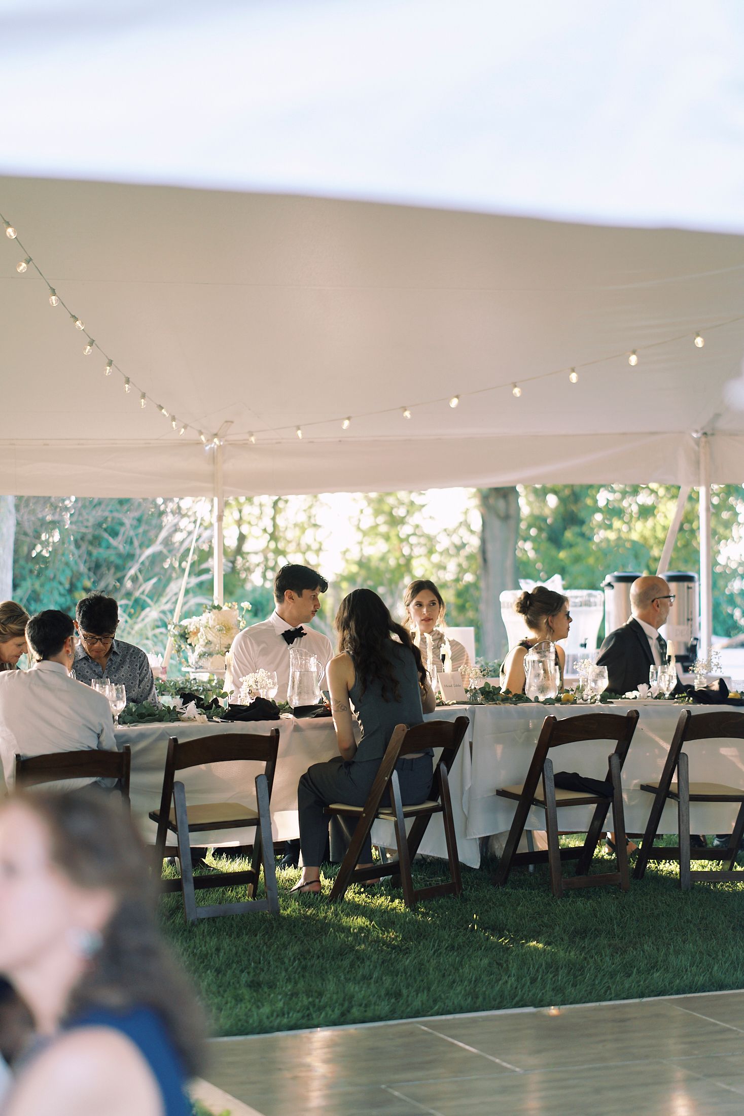 Guests seated at tables under an elegant white outdoor tent during a wedding reception with string lights overhead.