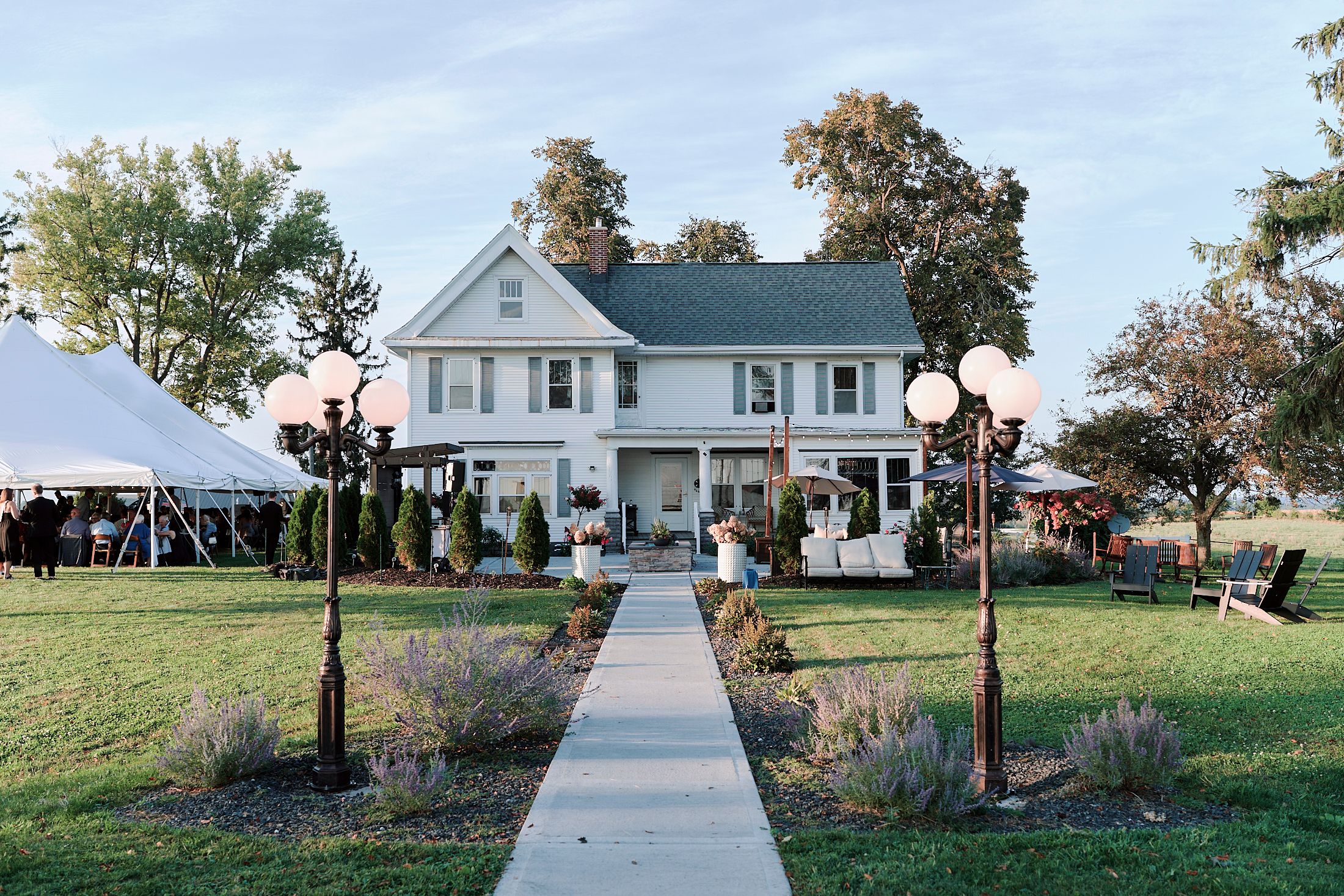 A classic white two-story farmhouse with a long walkway and decorative lamp posts leading to the entrance.