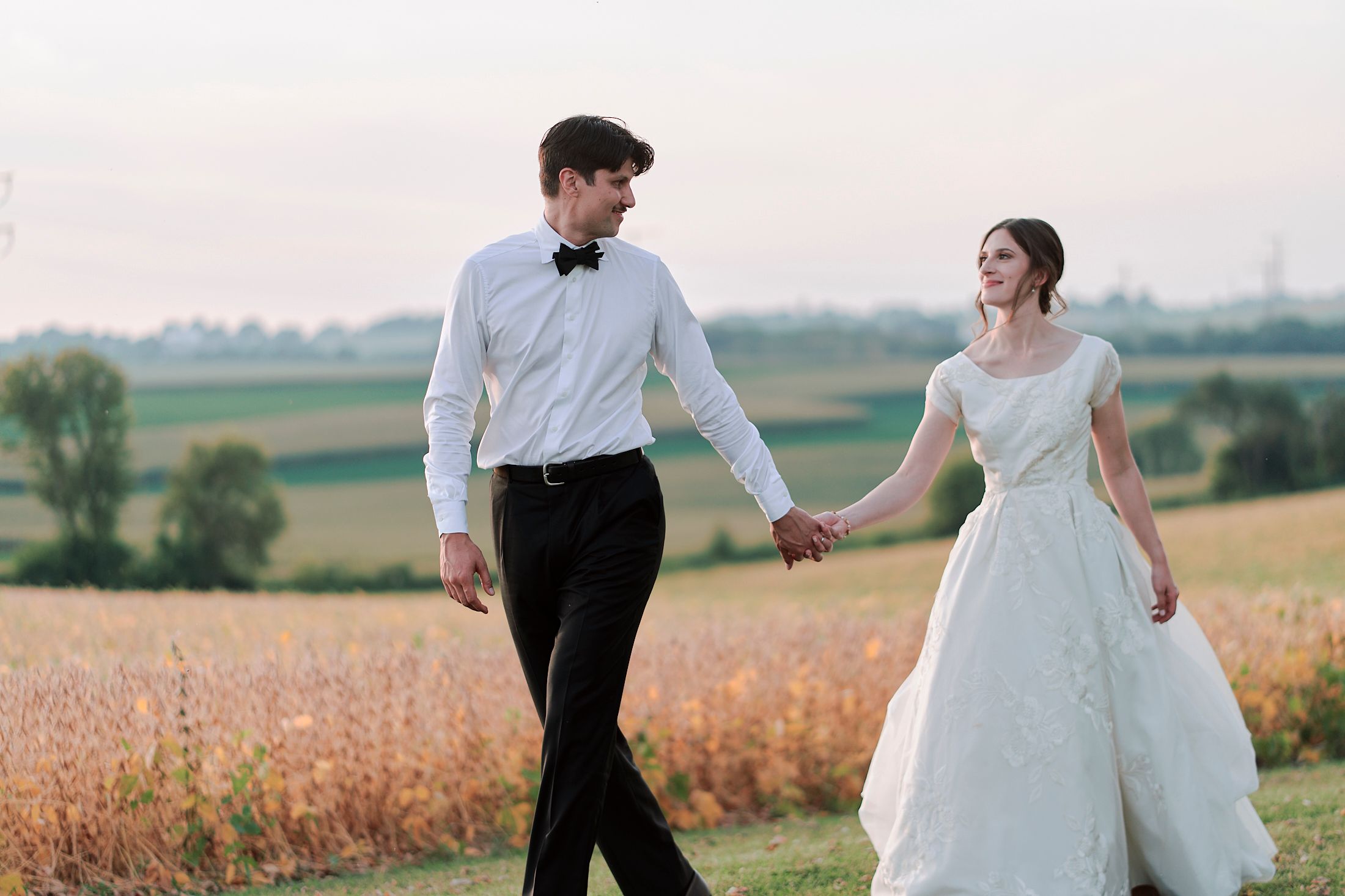 A couple walks hand in hand through a golden wheat field at sunset during their wedding celebration.