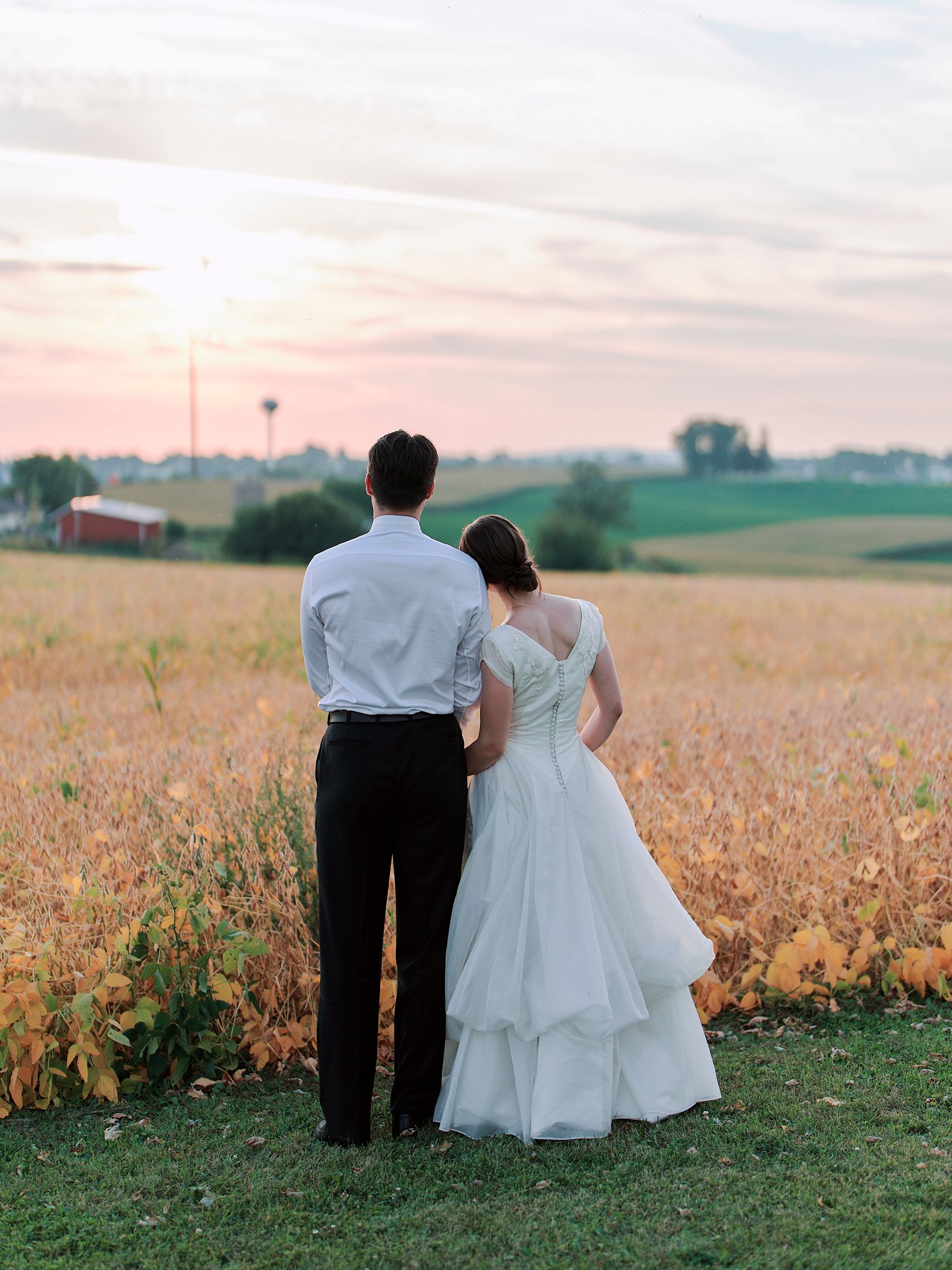 A couple in wedding attire stands together overlooking a scenic rural landscape at sunset.