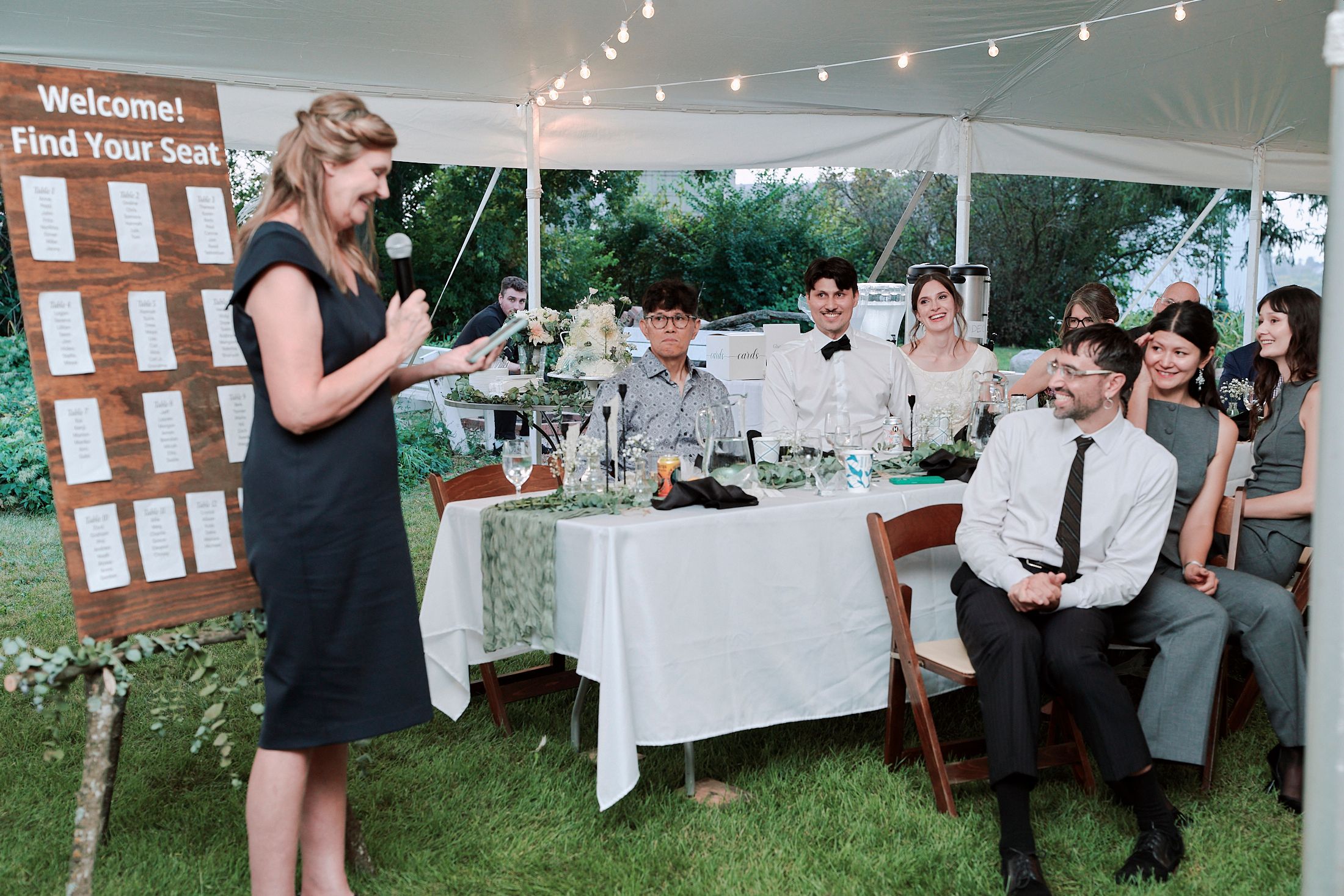 Wedding guests mingle and socialize at an outdoor reception with string lights and seating chart displays.