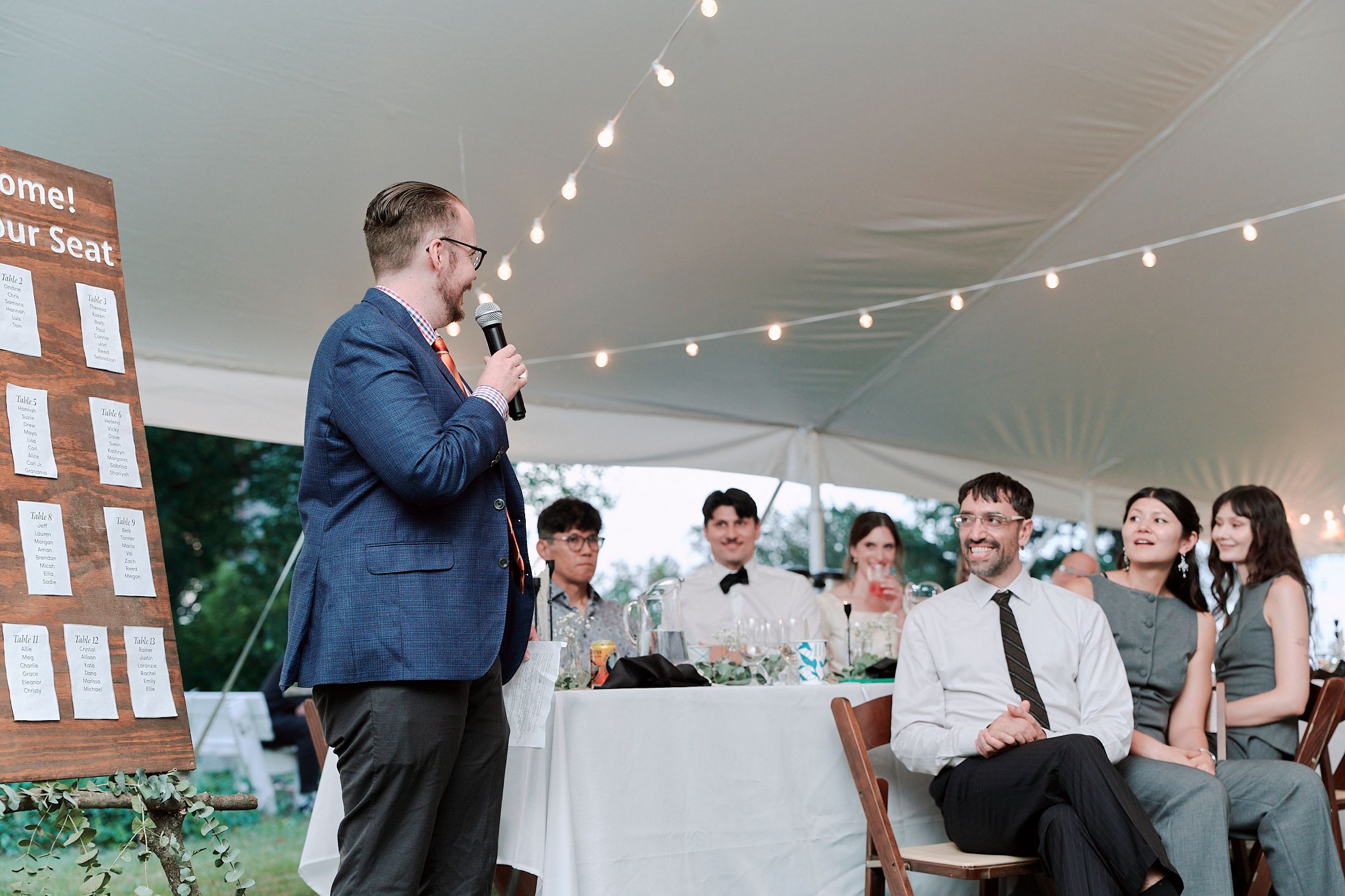 Wedding reception guests mingle and socialize under a white tent decorated with string lights and paper lanterns.
