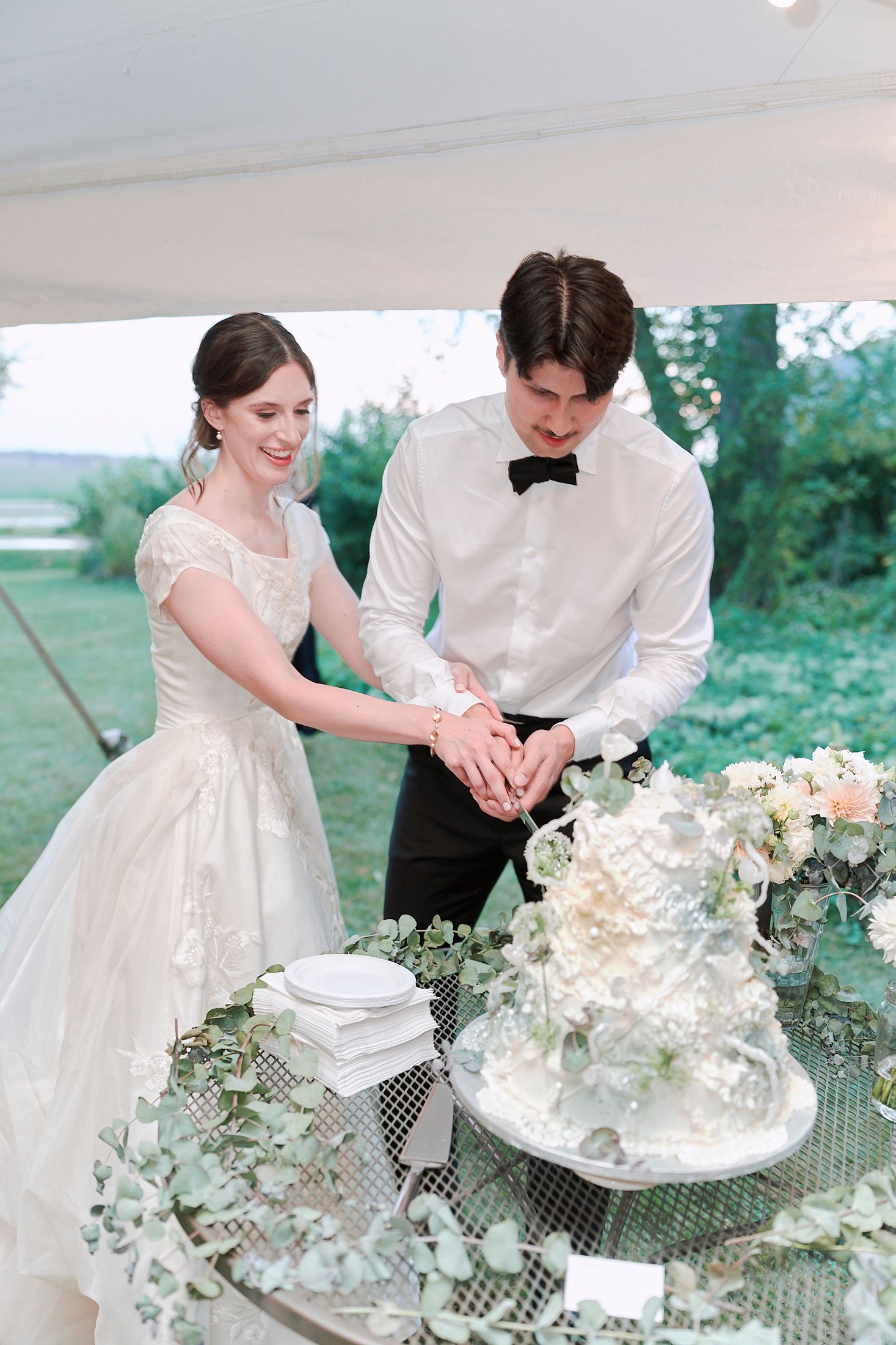 Newlywed couple cuts into an elegant white wedding cake decorated with fresh flowers on a garden table.